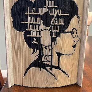 May include: A distinctive book art piece depicting a person in silhouette with glasses and a bun, with an intricate interior design. The silhouette is cut from book pages, creating a striking black and beige contrast.