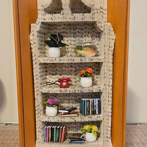 Peut inclure: Une sculpture de bibliothèque unique créée à partir d'un livre recyclé. L'intérieur présente des étagères avec des livres miniatures, des plantes, un téléphone rotatif rouge et un bocal à poissons. Les pages du livre sont pliées pour créer les étagères.