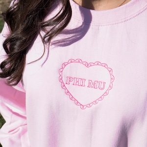 May include: A light pink sweatshirt with a heart-shaped embroidered design. The heart features the text "PHI MU" in a matching pink color, surrounded by a scalloped border. The sweatshirt is a solid color.