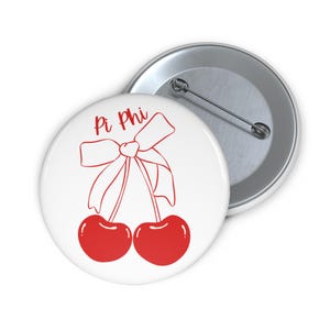 May include: White button with red cherries and a red bow with the text "Pi Phi" in red.