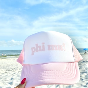 May include: A pink and white trucker hat with the text "phi mu!" in a light pink font. The hat has a pink brim and mesh sides. The background is a beach scene with blue sky and water.