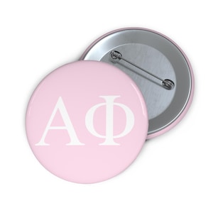 May include: A round pin-back button featuring a light pink background with the white Greek letters Alpha and Phi printed in the center. The back of the button is metallic silver and includes a horizontal pin mechanism for attachment to clothing.