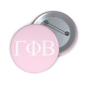 May include: A round pinback button featuring a light pink background with the white Greek letters Gamma Phi Beta printed in the center. The back of the button is metallic silver and includes a standard pin mechanism for attachment to clothing.