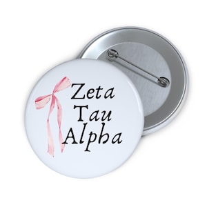 May include: White button with a pink ribbon and the text "Zeta Tau Alpha" in black.