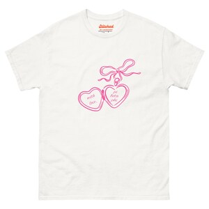Könnte beinhalten: Weißes T-Shirt mit einem rosa herzförmigen Medaillon-Design. Das Medaillon hat die Worte "with love" und "pi beta phi" darin. Über dem Medaillon befindet sich eine rosa Schleife. Das Shirt hat oben den Text "Stitched".