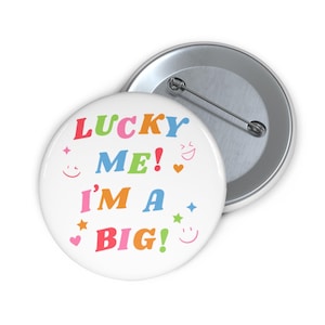 May include: A white button pin with the text "LUCKY ME! I'M A BIG!" in colorful letters. The pin has a metal backing and a safety pin closure. Decorative elements include hearts, stars, and smiley faces.
