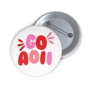May include: Two white round buttons with a pin back. One button has the words "GO AOU" in red and pink bubble letters. The other button is silver. The text is surrounded by red and pink starbursts.