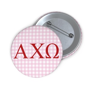 May include: Two round buttons. The front button has a pink and white gingham pattern with the Greek letters AXΩ in red. The back of the button is silver with a pin. The second button is partially visible.
