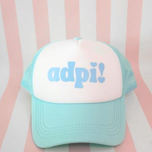 May include: A light blue and white trucker hat with the word "adpi!" in light blue script. The hat has a curved brim and a mesh back. The background is a pink and white striped pattern.