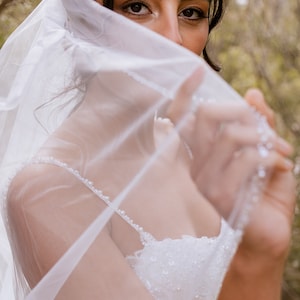 May include: A bride wearing a white wedding dress with a veil covering her face. The veil is decorated with small pearls. The bride is looking at the camera.