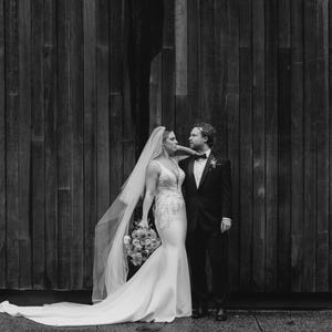 May include: A bride and groom pose for a wedding portrait in front of a wooden wall. The bride is wearing a white wedding dress with a long veil and a bouquet of flowers. The groom is wearing a black suit and a white shirt with a black bow tie.
