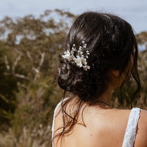 Könnte beinhalten: Eine Frau mit langen dunklen Haaren trägt ein weißes Kleid mit einem floralen Haarschmuck. Der Haarschmuck hat kleine weiße Blüten und perlmuttartige Akzente.