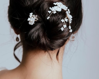 May include: A close-up of a dark-haired bun adorned with delicate white floral hairpins. The pins feature small, pearl-like beads and are arranged to add a touch of elegance to the hairstyle. The background is a soft, neutral color.