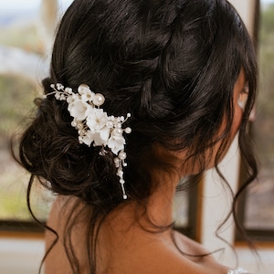 以下が含まれることがあります： 真珠のアクセントが付いた白い花のヘアコーム。コームは、緩いお団子にまとめられた女性の濃い茶色の髪につけられています。