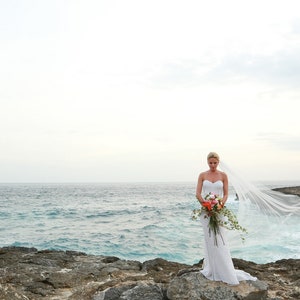 May include: A bride in a white wedding dress with a long veil stands on a rocky cliff overlooking the ocean. The veil is blowing in the wind, and the bride is holding a bouquet of flowers.