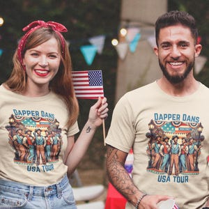 May include: Two people wearing beige t-shirts with a graphic print of a group of men in vintage clothing. The graphic includes the text "Dapper Dans USA Tour".