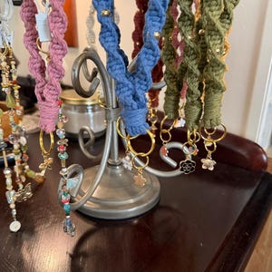 May include: A display of handmade macrame keychains in various colors, including pink, blue, and green. Each keychain features a spiral design, adorned with gold beads and charm pendants. The keychains are hanging on a decorative metal stand.