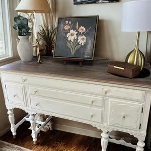 Puede incluir: Un aparador antiguo blanco y marrón con herrajes dorados. El aparador tiene cajones y puertas de armario. Una pintura floral enmarcada, una caja decorativa y dos lámparas están encima.