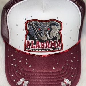 May include: A burgundy and white trucker hat featuring the text "ALABAMA CRIMSON TIDE" and an elephant logo. The hat is decorated with rhinestones and small white bows.
