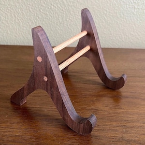 May include: A wooden stand with two angled supports and a crossbar. The stand is made of dark brown wood with a natural wood finish. The stand is designed to hold a plate or other flat object.