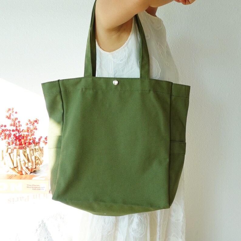 May include: Olive green canvas tote bag with two exterior pockets and a silver snap closure. The bag has two sturdy handles for carrying. The tote bag is a simple, yet stylish accessory.