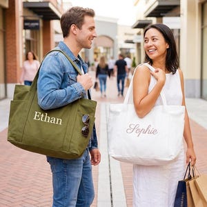 Puede incluir: Dos personas caminando en una zona comercial al aire libre, cada una llevando un bolso tote grande. Una bolsa es de color verde oliva con el nombre "Ethan" en blanco. La otra es blanca con el nombre "Sophie" en cursiva.