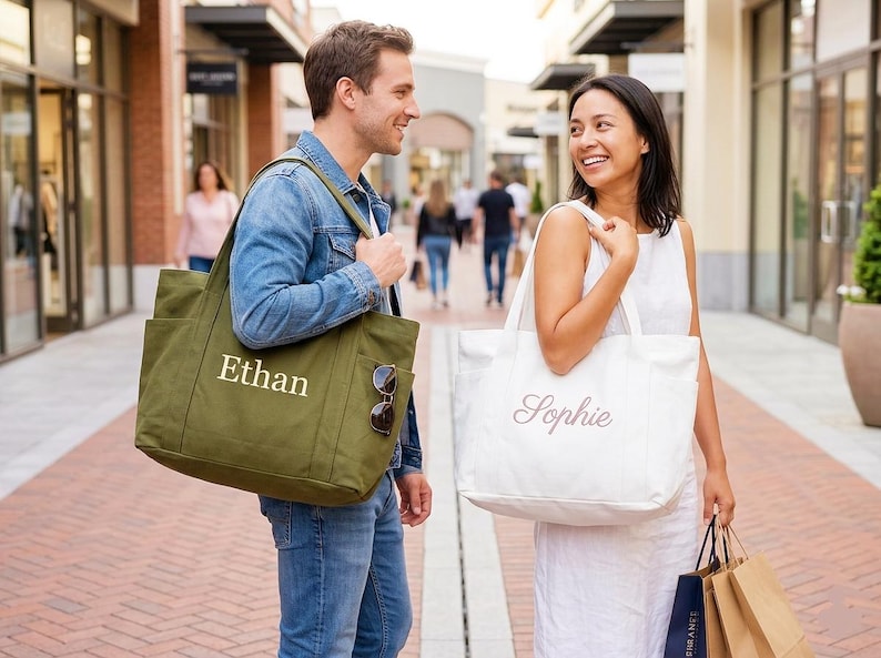 Puede incluir: Dos personas caminando en una zona comercial al aire libre, cada una llevando un bolso tote grande. Una bolsa es de color verde oliva con el nombre "Ethan" en blanco. La otra es blanca con el nombre "Sophie" en cursiva.
