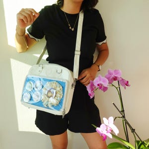 May include: White crossbody bag with a clear front pocket displaying several circular pins and a decorative item. The bag has a heart-shaped detail and a long, adjustable strap. The bag is worn over the shoulder.