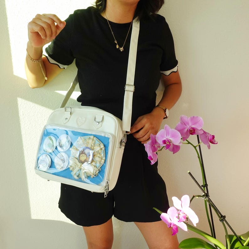May include: White crossbody bag with a clear front pocket displaying several circular pins and a decorative item. The bag has a heart-shaped detail and a long, adjustable strap. The bag is worn over the shoulder.