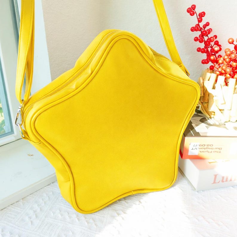 May include: A bright yellow star-shaped shoulder bag with a zipper closure and a long strap. The bag is made of a smooth material and has a rounded shape. The bag is sitting on a white surface.