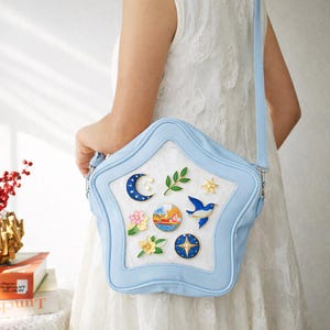 May include: A light blue star-shaped crossbody bag with a white center, adorned with various pins including a crescent moon, a bird, and flowers. The bag has a long strap and silver hardware.