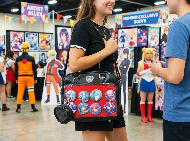 May include: Black shoulder bag with a red interior, displaying several anime character buttons. The bag has a small, heart-shaped charm and a small, round coin purse attached. The background shows anime-themed posters and cosplayers.