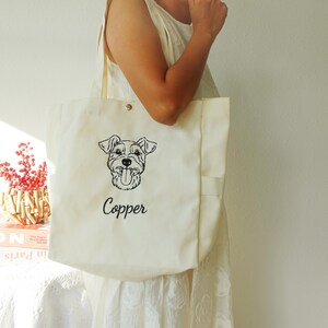 May include: Cream-colored canvas tote bag featuring a black line drawing of a dog's face with its tongue out. The name "Copper" is written in cursive below the dog illustration. The bag has a snap closure and a pocket on the side.
