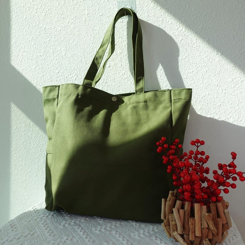 May include: Olive green canvas tote bag with long handles, perfect for carrying groceries or everyday essentials. The bag has a simple, minimalist design with a single snap closure. A decorative wooden vase with red berries sits nearby.