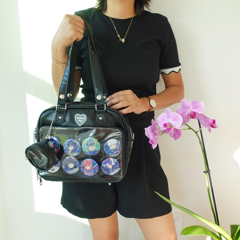 May include: Black satchel bag with a clear front pocket displaying colorful character pins. The bag has a small matching coin purse attached. A heart-shaped emblem is visible on the front. The bag is held by a person wearing a black outfit.