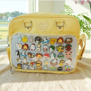 May include: A yellow satchel bag with a clear front panel displaying a collection of colorful enamel pins. The pins feature various cartoon characters and designs. The bag has silver hardware and a shoulder strap.