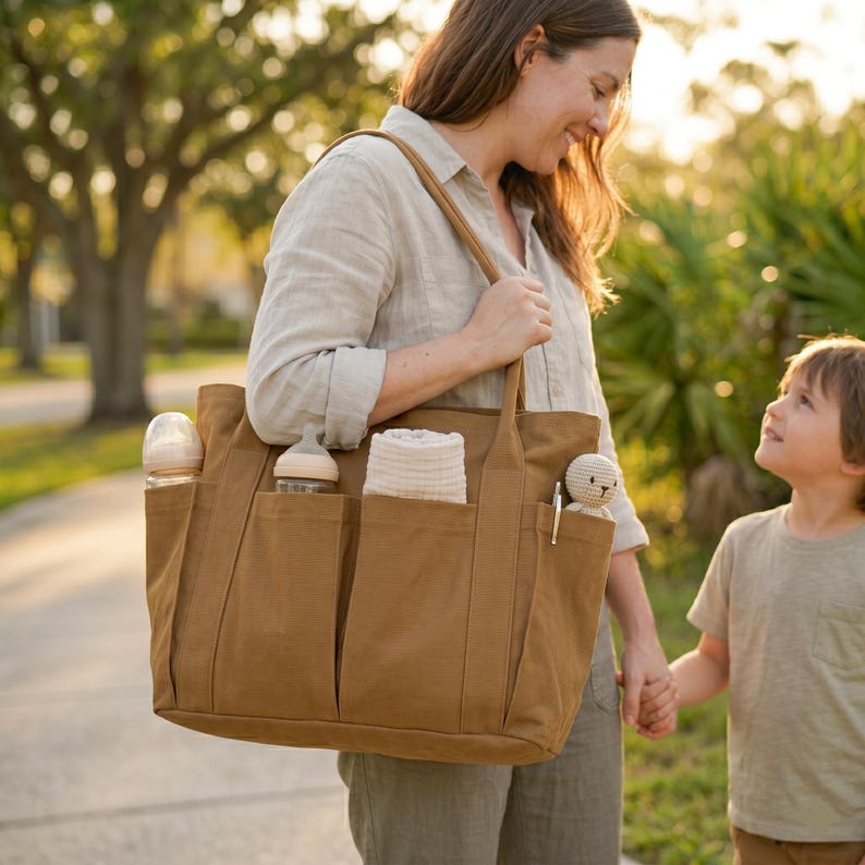 Puede incluir: Un bolso tote marr&oacute;n grande con m&uacute;ltiples bolsillos exteriores, que contiene biberones, un pa&ntilde;o enrollado y un peque&ntilde;o peluche. El bolso lo lleva una persona, y su contenido sugiere que es un bolso para pa&ntilde;ales.