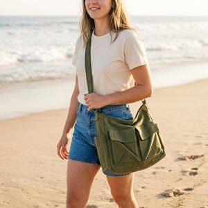 May include: An olive green canvas shoulder bag with multiple front pockets and a long strap. The bag is carried by a person walking on a sandy beach. The person is wearing denim shorts and a cream-colored t-shirt.