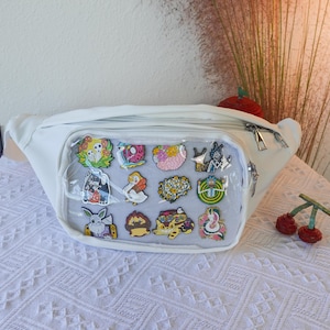 May include: White fanny pack with a clear plastic front panel displaying a variety of colorful enamel pins. The pins feature cartoon characters, animals, and abstract designs.