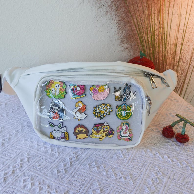 May include: White fanny pack with a clear plastic front panel displaying a variety of colorful enamel pins. The pins feature cartoon characters, animals, and abstract designs.