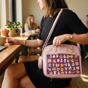 May include: A light pink crossbody bag with a clear front panel displaying a collection of colorful character pins. The bag has a matching small side pouch and a silver chain strap. The bag is held by a person wearing a black dress.