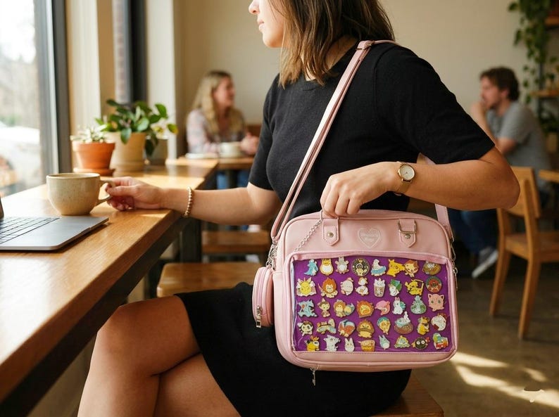 May include: A light pink crossbody bag with a clear front panel displaying a collection of colorful character pins. The bag has a matching small side pouch and a silver chain strap. The bag is held by a person wearing a black dress.