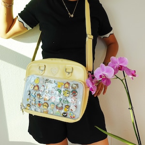 May include: A pale yellow shoulder bag with a clear front panel filled with colorful character pins. The bag has a heart-shaped detail and silver-colored hardware. The bag's strap is also pale yellow.