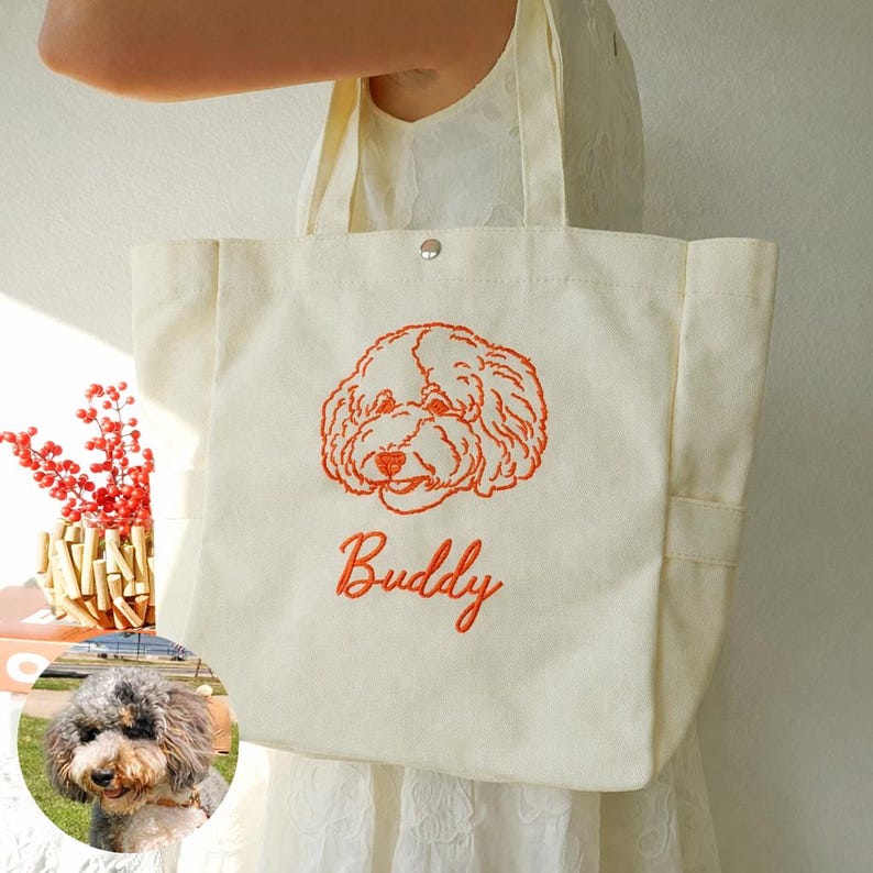 May include: Cream-colored tote bag with an embroidered orange dog face and the name "Buddy" in orange script. The bag has two handles and a snap closure. A small photo of a dog is in the lower left corner.