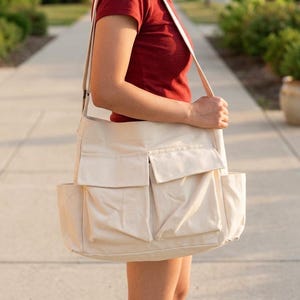 May include: A cream-colored canvas tote bag with multiple exterior pockets and a shoulder strap. The bag is carried by a person wearing a red shirt and shorts, walking on a sidewalk.
