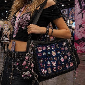 Puede incluir: Un bolso negro con un panel frontal transparente lleno de coloridos pines de estilo anime. El bolso est&aacute; adornado con cadenas y abalorios. La persona lleva un top negro con detalles en rosa, un cintur&oacute;n y un cord&oacute;n.