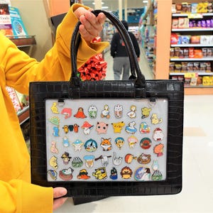 May include: Black tote bag with a faux crocodile skin texture, holding a clear panel filled with colorful enamel pins. The pins feature various cartoon characters, food items, and other designs. The bag has black handles.
