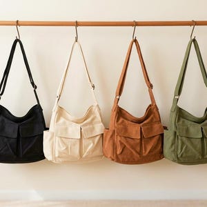May include: Four canvas messenger bags in black, cream, brown, and olive green hang from a wooden rod. Each bag has a front flap pocket and an adjustable shoulder strap. The bags are displayed against a neutral background.