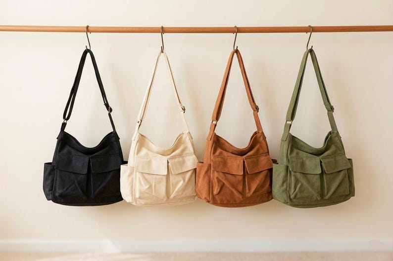 May include: Four canvas messenger bags in black, cream, brown, and olive green hang from a wooden rod. Each bag has a front flap pocket and an adjustable shoulder strap. The bags are displayed against a neutral background.