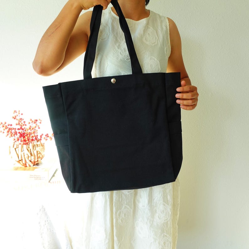 May include: A black tote bag with long handles and a silver snap closure. The bag has a rectangular shape with side pockets and is made of a sturdy fabric. The tote bag is held up by a person.